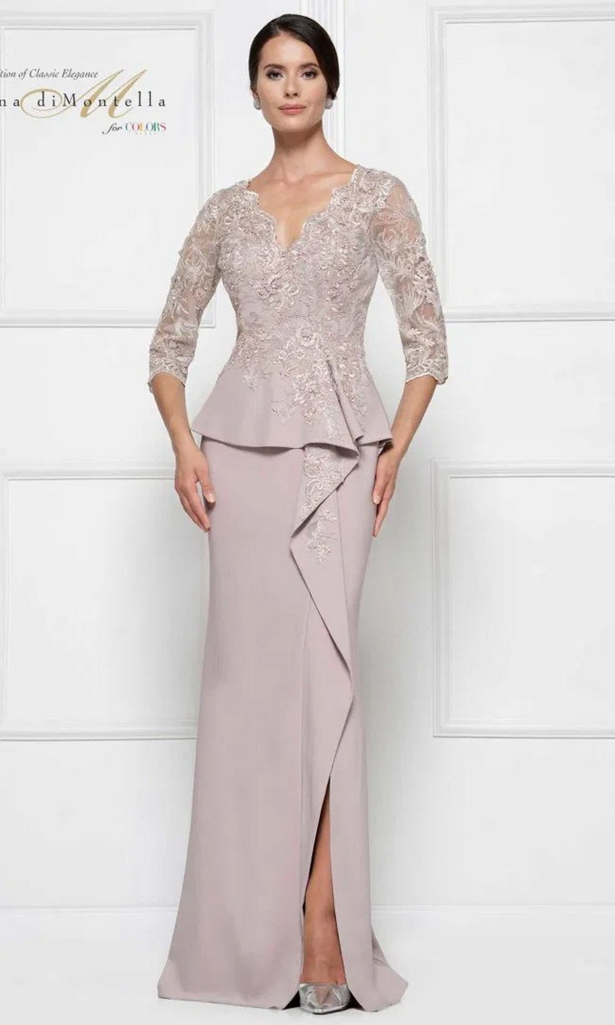 A woman stands against a white paneled wall wearing the Rina Di Montella RD2685 long formal dress in taupe, featuring lace embroidered sleeves, a V-neckline, peplum waist, and a floor-length sheath skirt.